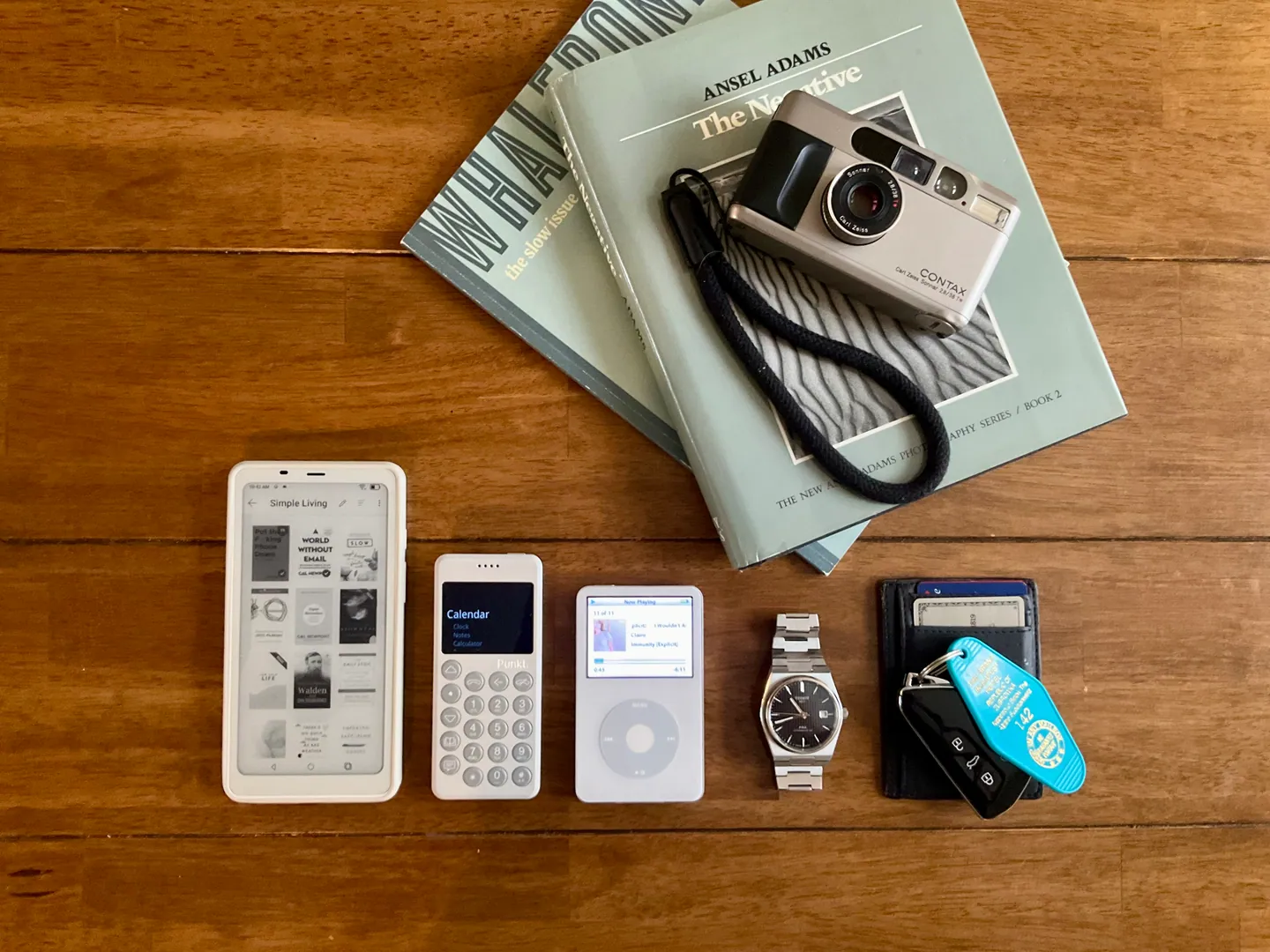 An old everyday carry laid out on a wooden table: an e-ink phone, a Punkt keypad phone, an iPod, a steel watch, a wallet with keys, a Contax film camera on two photography books